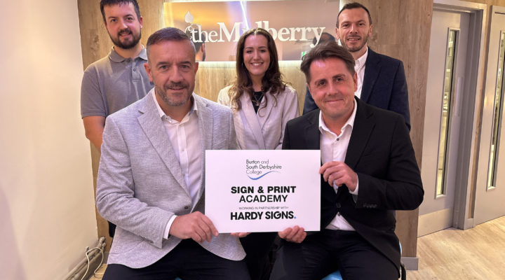 Five smartly dressed people smiling at the camera. The two men seated at the front of the group are holding a white sign saying "Burton and South Derbyshire College, Sign and Print Academy , working in partnership with Hardy Signs."